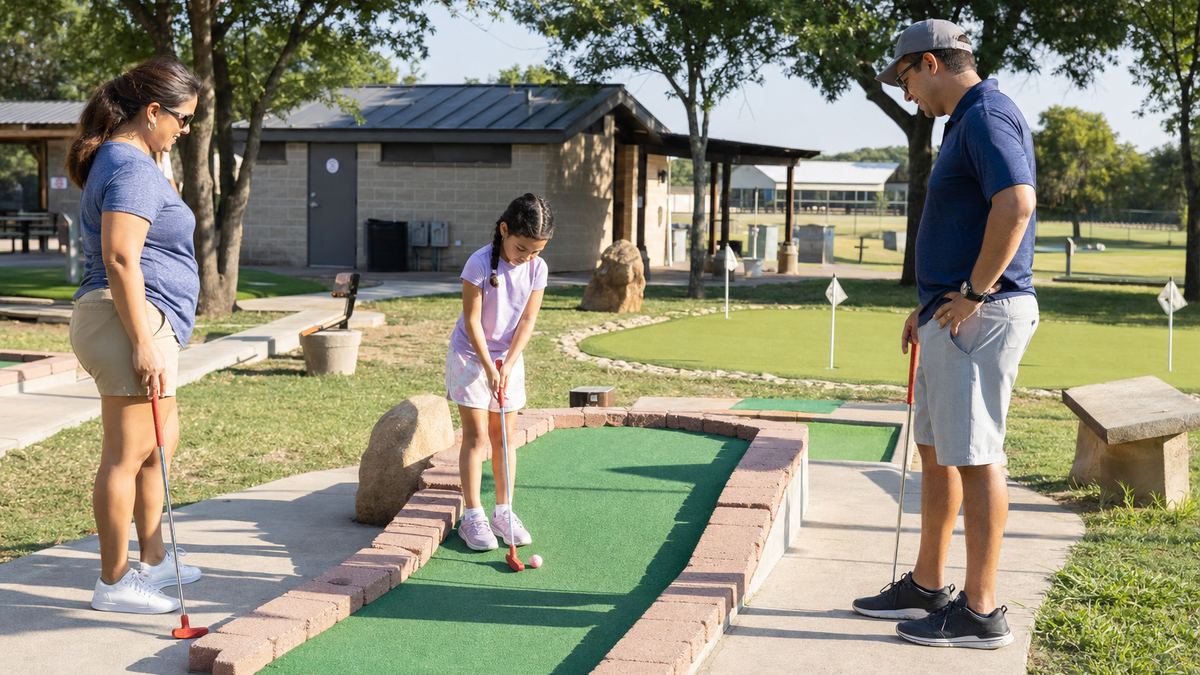 A family playing a hole on an outdoor miniature golf course with green turf and low borders at Mary Jo Peckham Park in Katy, Texas